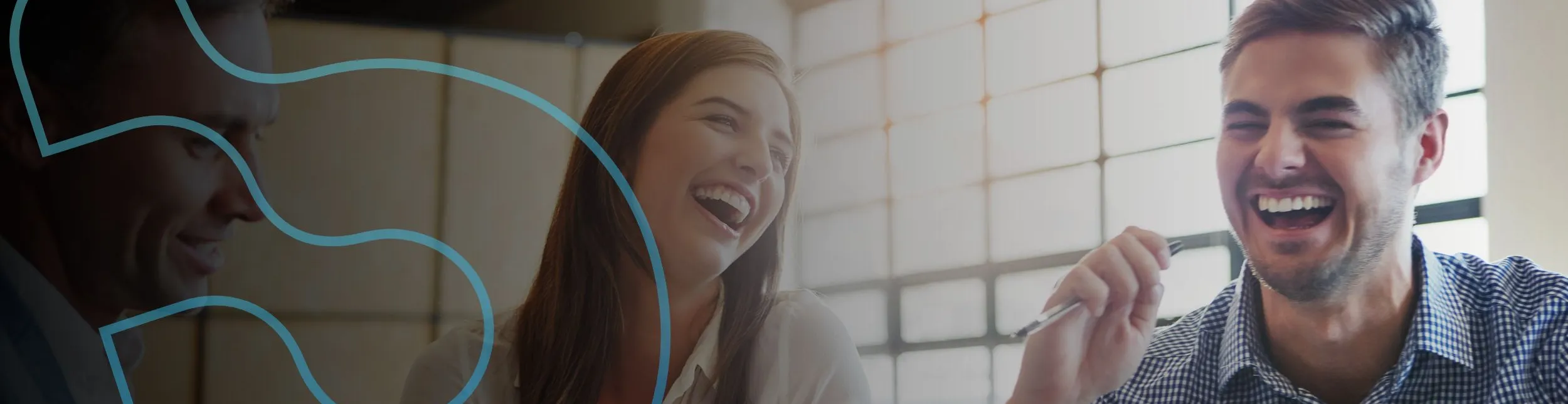 Dois colegas de trabalho sentados próximos a uma janela. Uma mulher ri enquanto conversa com um homem que também sorri segurando uma caneta. A imagem transmite leveza e descontração.