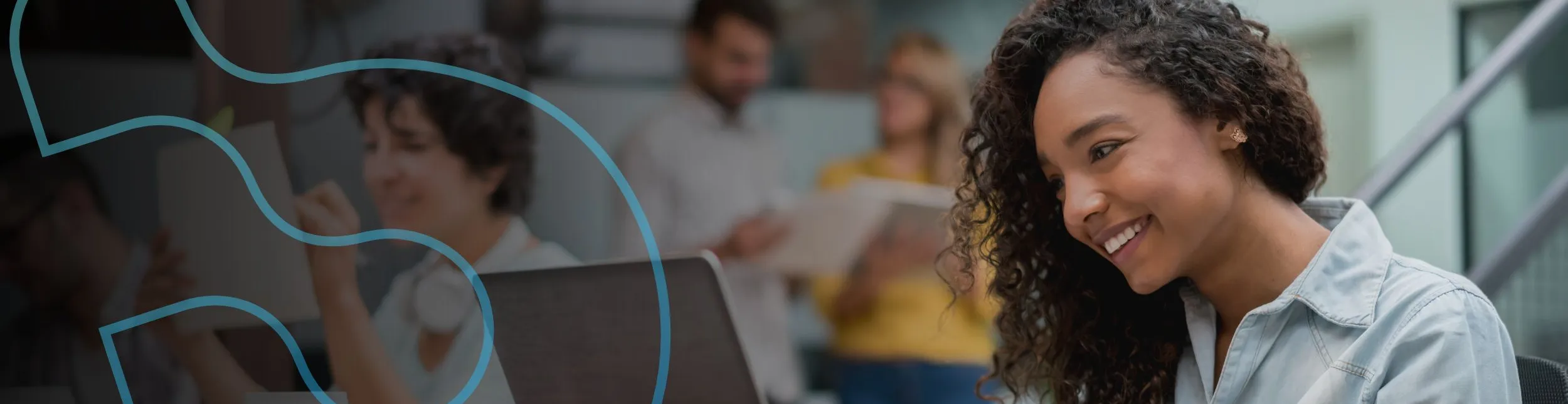 Mulher jovem de cabelos cacheados sorrindo enquanto olha para a tela de um notebook. Atrás dela, aparecem colegas desfocados trabalhando em um escritório moderno.