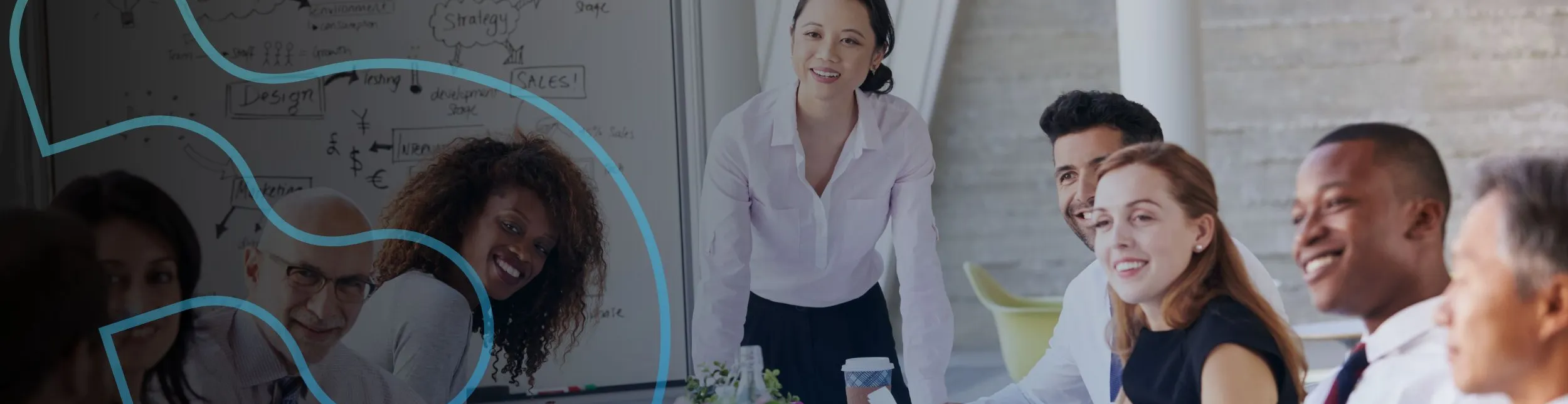 Grupo diverso de profissionais sentados ao redor de uma mesa em uma sala de reunião iluminada. Uma mulher em pé, ao centro, sorri enquanto conversa com o time. Todos parecem engajados e colaborando.