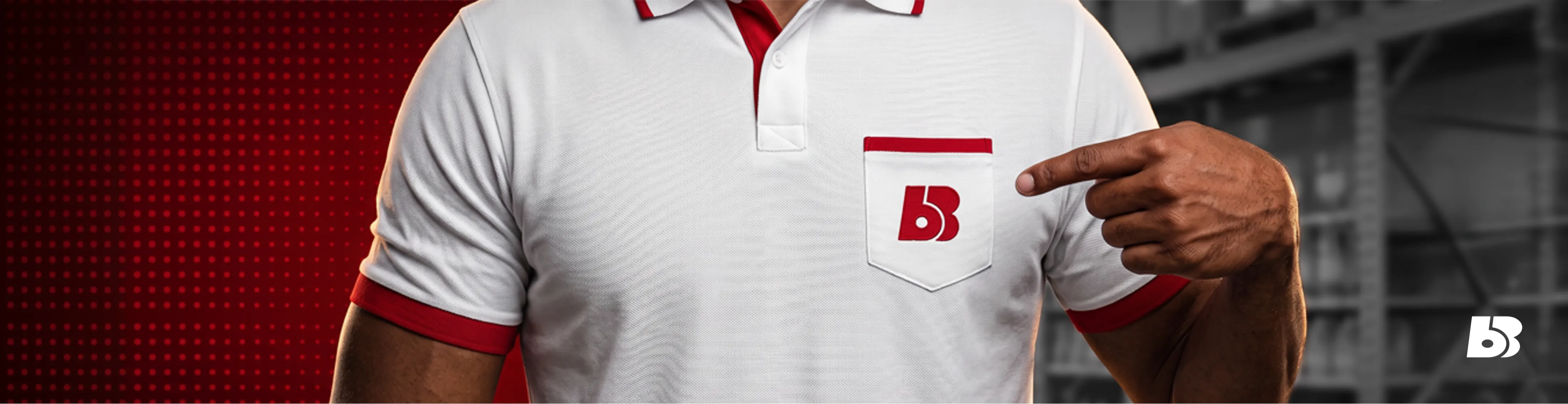 Uma fotografia em "zoom de câmera" de um homem com pele parda, cortada do peito para baixo, vestindo uma camisa polo branca. A camisa tem um bolso no peito direito com o logotipo vermelho e branco 'BB' do 'Big Bom'. A camisa tem acabamento vermelho na gola, nas mangas e na aba do peitilho. O homem aponta o dedo indicador direito diretamente para o logotipo. O fundo é dividido, com um painel vermelho sólido com uma grade de pontos à esquerda e uma vista cinza desfocada de prateleiras de armazenamento de armazém à direita.