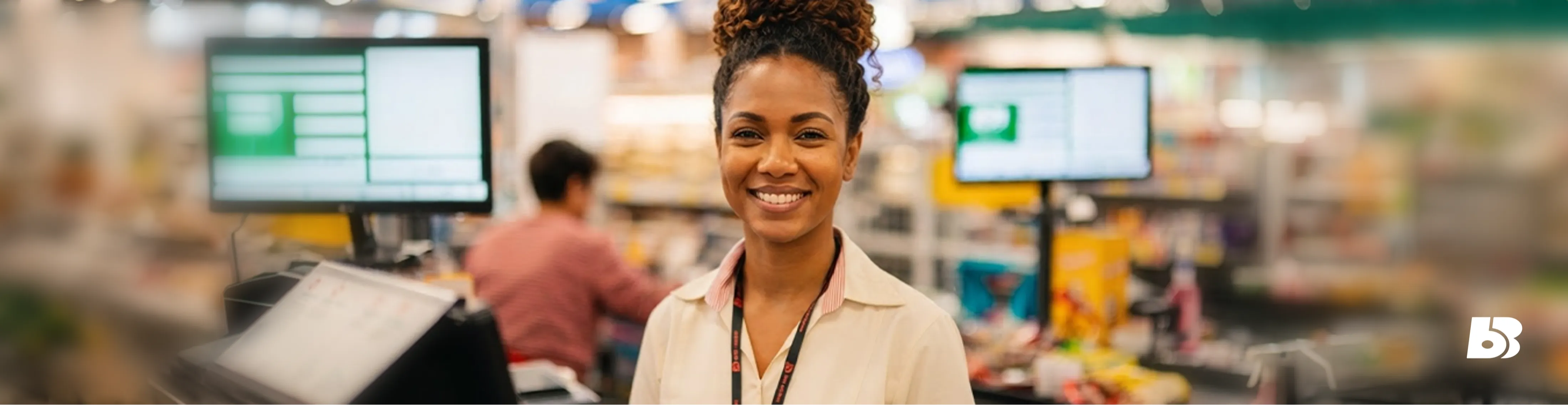 Fotografia em formato de banner horizontal com o plano médio de uma colaboradora mulher do Big Bom Supermercados. Ela está posicionada ao centro, sorrindo de forma simpática e acolhedora para a câmera. É uma mulher negra, de pele clara, com cabelos cacheados em tons de castanho, presos com um coque na parte superior da cabeça. Ela veste o uniforme da empresa: uma camisa social de manga curta na cor off-white, com um detalhe vermelho na gola e o logotipo bordado discretamente no peito, além de um cordão de crachá preto com escritas em vermelho. Ela está na área dos caixas de supermercado (frente de loja), com os equipamentos de PDV (Ponto de Venda) e monitores de computador visíveis ao fundo de forma suavemente desfocada. No canto inferior direito, há a aplicação do ícone 'BB' do logotipo da empresa na cor branca.