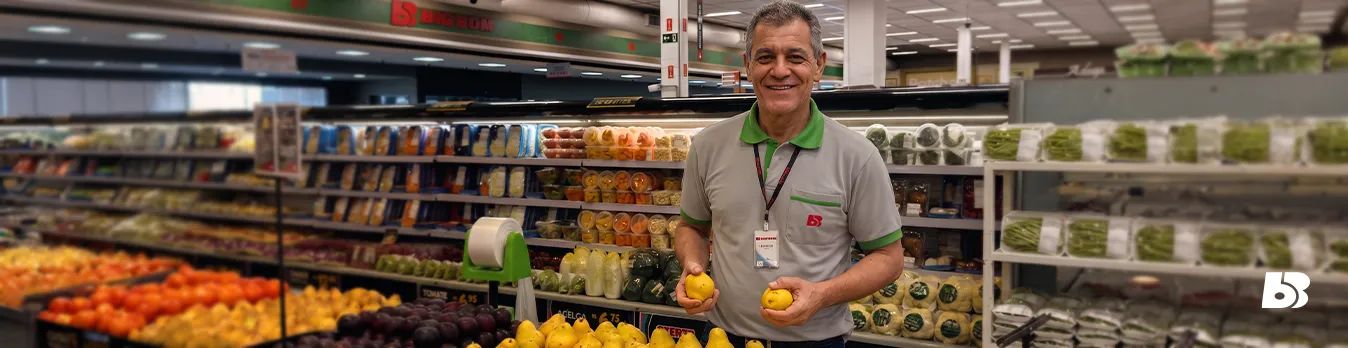 Colaborador do hortifrúti
A quarta fotografia retrata um colaborador do setor de hortifrúti, posicionado ao centro da imagem. Ele veste uniforme cinza com gola verde e crachá, sorrindo enquanto segura duas frutas amarelas, uma em cada mão. Ao seu redor há uma grande variedade de frutas, legumes e verduras expostos em bancas refrigeradas e prateleiras. O ambiente é amplo, muito iluminado e evidencia organização e abundância dos produtos.