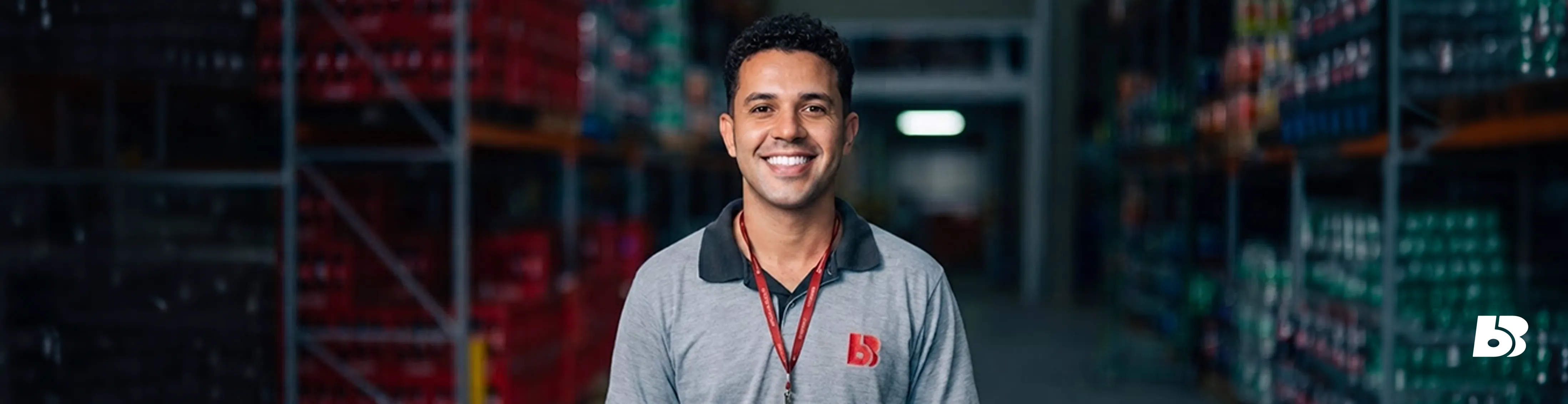 Fotografia em formato de banner horizontal com o plano médio de um colaborador homem do Big Bom Supermercados. Ele está centralizado na imagem, sorrindo de forma aberta e confiante para a câmera. É um homem de pele parda, com cabelos pretos curtos e crespos. Veste o uniforme da empresa: uma camisa polo cinza com gola cinza-escura, o logotipo vermelho bordado no lado esquerdo do peito e um cordão de crachá vermelho no pescoço. O fundo retrata o ambiente de um estoque ou depósito de mercadorias, com estruturas de prateleiras industriais repletas de caixas e produtos, de forma suavemente desfocada. No canto inferior direito, há a aplicação do ícone 'BB' do logotipo da empresa na cor branca.