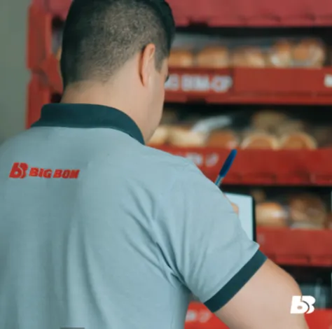 Fotografia quadrada mostrando o plano médio de um colaborador homem do Big Bom Supermercados, visto de costas e de perfil de três quartos. Ele tem pele clara e cabelos pretos bem curtos. O profissional veste o uniforme da empresa: uma camisa polo cinza-claro com a gola e a borda das mangas em cinza-escuro. Nas costas da camisa, na altura do ombro esquerdo, destaca-se o logotipo bordado em vermelho com o ícone 'BB' e o nome 'BIG BOM'. Ele aparece segurando uma caneta azul e fazendo anotações em uma prancheta. À sua frente, há uma pilha de caixas plásticas organizadoras vermelhas repletas de produtos de panificação embalados em saquinhos transparentes, como pães. No canto inferior direito, há a aplicação do ícone 'BB' do logotipo na cor branca.