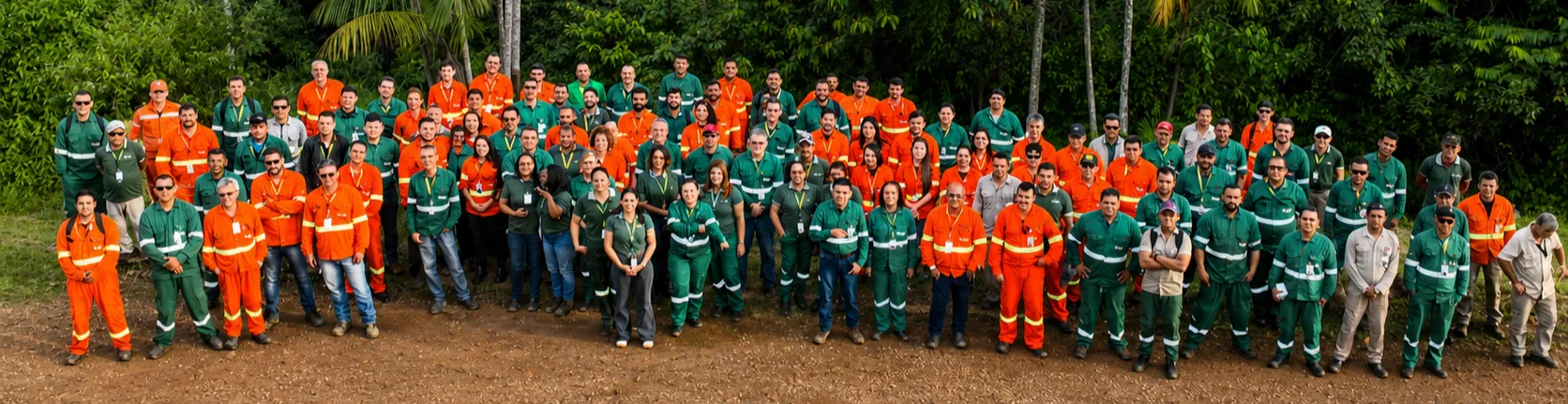 Foto panorâmica de um grande grupo de profissionais da Amapá Minerals reunido ao ar livre. As pessoas usam uniformes verdes, laranjas e cinza, com faixas refletivas, e estão posicionadas em várias fileiras sobre uma área de terra. Ao fundo, há vegetação densa, com árvores e palmeiras, formando uma paisagem de floresta.