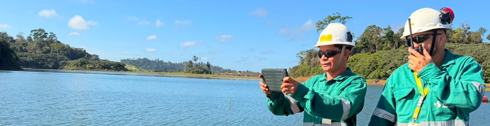 Dois profissionais da Amapá Minerals, usando uniformes verdes, capacetes e óculos de proteção, realizam uma atividade de monitoramento às margens de um reservatório. Um deles segura um tablet, enquanto o outro utiliza um rádio comunicador. Ao fundo, há vegetação e céu azul.