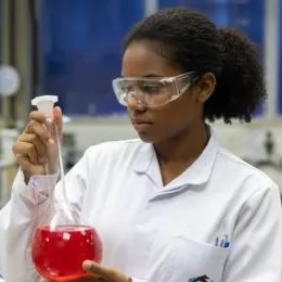 A imagem mostra um ambiente de laboratório. No primeiro plano, há uma pessoa vestindo um jaleco branco e óculos de proteção. A pessoa segura um frasco volumétrico transparente contendo um líquido vermelho intenso. A mão está posicionada junto ao gargalo do frasco, segurando também uma pipeta volumétrica ou instrumento semelhante, indicando manipulação de substâncias químicas.
Ao fundo, é possível ver um ambiente típico de laboratório: superfícies de trabalho, equipamentos desfocados e um painel azul que compõe o cenário.