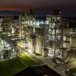 A imagem mostra um complexo industrial iluminado à noite. A estrutura ocupa grande parte da cena e é composta por diversos prédios, passarelas metálicas, tubulações, torres e plataformas. As luzes artificiais, distribuídas por toda a instalação, criam um destaque forte contra o céu noturno escuro.
No lado esquerdo, há áreas com gramado bem iluminado e trechos de tubulações horizontais. No centro e à direita, sobressaem edifícios industriais maiores, com vários andares, janelas iluminadas e estruturas verticais típicas de plantas de processamento ou produção química.
Ao fundo, é possível ver um horizonte com relevo montanhoso levemente visível, sugerindo que a instalação está situada em uma área mais ampla e aberta.