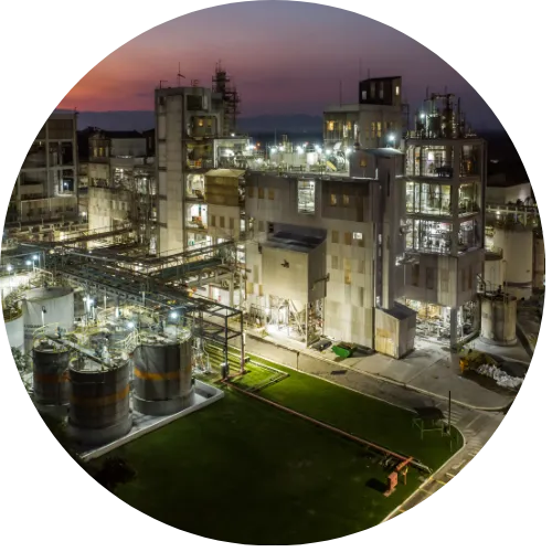 A imagem mostra um complexo industrial iluminado à noite. A estrutura ocupa grande parte da cena e é composta por diversos prédios, passarelas metálicas, tubulações, torres e plataformas. As luzes artificiais, distribuídas por toda a instalação, criam um destaque forte contra o céu noturno escuro.
No lado esquerdo, há áreas com gramado bem iluminado e trechos de tubulações horizontais. No centro e à direita, sobressaem edifícios industriais maiores, com vários andares, janelas iluminadas e estruturas verticais típicas de plantas de processamento ou produção química.
Ao fundo, é possível ver um horizonte com relevo montanhoso levemente visível, sugerindo que a instalação está situada em uma área mais ampla e aberta.