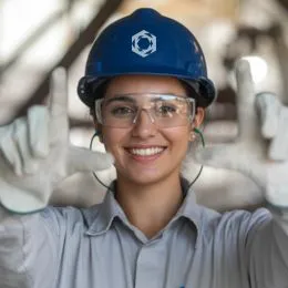 A imagem mostra uma pessoa vestindo um capacete azul com um símbolo branco na parte frontal. A pessoa está usando óculos de proteção e luvas brancas. As mãos estão levantadas à altura da cabeça, formando um retângulo ou moldura com os dedos. A roupa é um uniforme de cor clara, típico de ambiente industrial.
O fundo apresenta uma estrutura metálica desfocada, sugerindo um cenário de instalação industrial ou operacional.