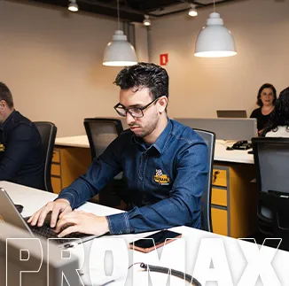 Homem utilizando óculos e camisa corporativa trabalha em um notebook em um ambiente de escritório. Sobre a mesa há um celular, um caderno e materiais de trabalho. Ao fundo, outras estações de trabalho e pessoas aparecem em um espaço compartilhado, iluminado por luminárias suspensas. A imagem representa atividades administrativas, tecnologia, colaboração e o ambiente corporativo da empresa. A marca "PROMAX" aparece em destaque na parte inferior da foto.