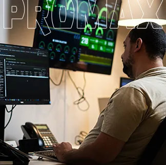 Homem sentado em uma estação de monitoramento que acompanha informações em telas de computador. Os monitores exibem gráficos, indicadores e dados operacionais em tempo real. Sobre a mesa há equipamentos de escritório, incluindo telefone e teclado. O ambiente remete a uma sala do departamento de tecnologia da informação. A marca "PROMAX" aparece em destaque na parte superior da foto.