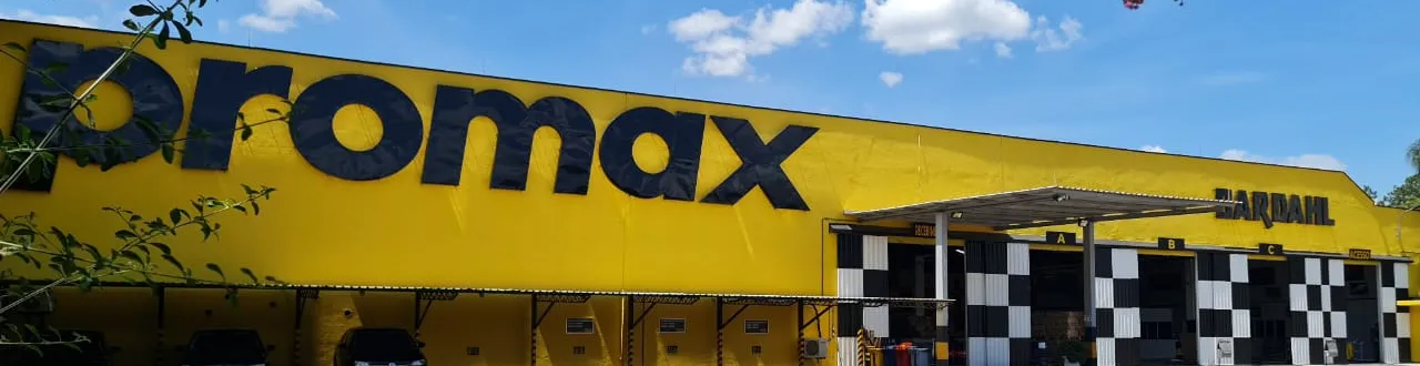 A imagem apresenta a fachada da Promax Bardahl, destacando os elementos visuais que compõem a identidade da empresa. O amarelo predominante da estrutura, combinado ao azul do logotipo Promax e aos detalhes quadriculados característicos da marca Bardahl, reforça atributos como energia, confiança, movimento e desempenho. A fotografia simboliza a presença da organização, sua trajetória de evolução contínua e o compromisso em mover pessoas, negócios e resultados.