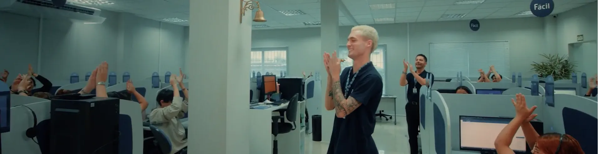 Escritório estilo call center com colaboradores em seus postos de trabalho aplaudindo e comemorando. No centro, um homem em pé sorri e bate palmas ao lado de uma coluna com um sino dourado, enquanto outras pessoas levantam as mãos em celebração. O ambiente é claro, organizado e transmite clima de reconhecimento e conquista.