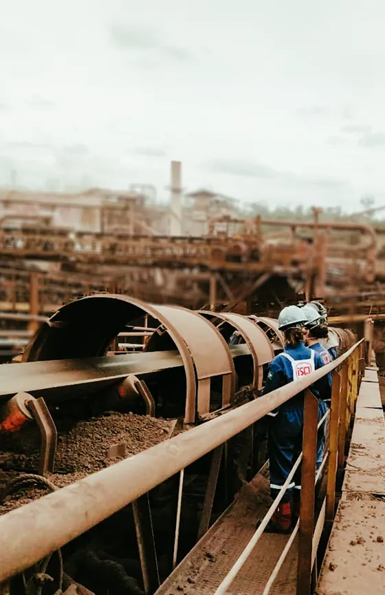 A imagem mostra o interior de uma área industrial. Em primeiro plano, há um transportador de correia carregando material escuro e granulado. A estrutura metálica ao redor está coberta de poeira e desgaste típico de ambientes industriais pesados.

À direita, sobre uma passarela metálica com corrimão, há três trabalhadoras ISQ usando equipamentos de proteção individual, incluindo macacões azuis, capacetes de segurança e botas. Elas estão de costas, observando o funcionamento do equipamento. Ao fundo, a paisagem é composta por diversas tubulações, estruturas metálicas e instalações industriais mais distantes, parcialmente desfocadas. O céu está claro, com nuvens finas.