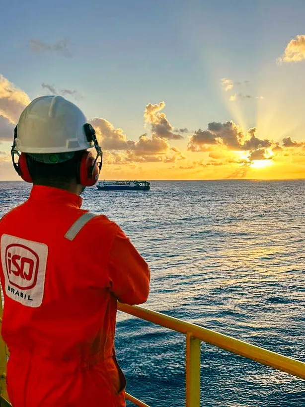Um trabalhador ISQ está em uma plataforma marítima, olhando para o mar durante o pôr do sol. Ele usa macacão laranja, capacete branco e protetores auriculares. Está de costas, apoiado no corrimão amarelo da estrutura. O sol se põe no horizonte, iluminando as nuvens com tons de dourado e laranja. Ao fundo, há uma embarcação distante sobre a água. A cena transmite tranquilidade e contemplação em ambiente industrial offshore.