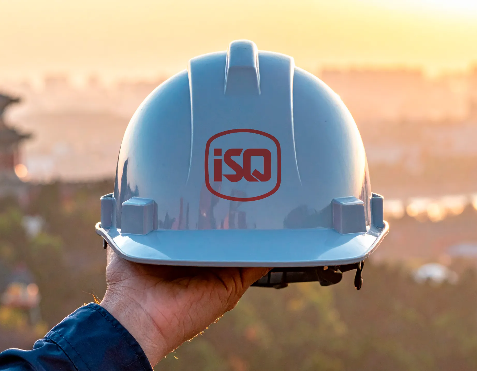 A imagem mostra uma mão segurando um capacete de segurança azul-claro com o logotipo vermelho do ISQ na frente. O fundo está desfocado e revela uma paisagem ao pôr do sol, com tons alaranjados e dourados iluminando a cena. Ao longe, aparecem construções e uma arquitetura tradicional, sugerindo um ambiente urbano com elementos culturais asiáticos. O foco está totalmente no capacete, simbolizando segurança, engenharia e atuação global.