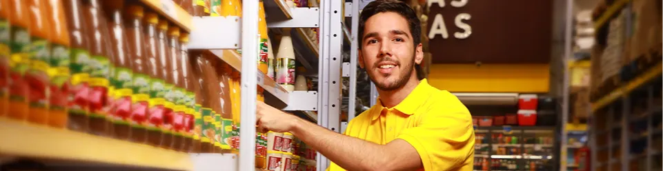 Funcionário sorridente, vestindo camisa amarela, organizando produtos em uma prateleira dentro da loja, cercado por embalagens coloridas.