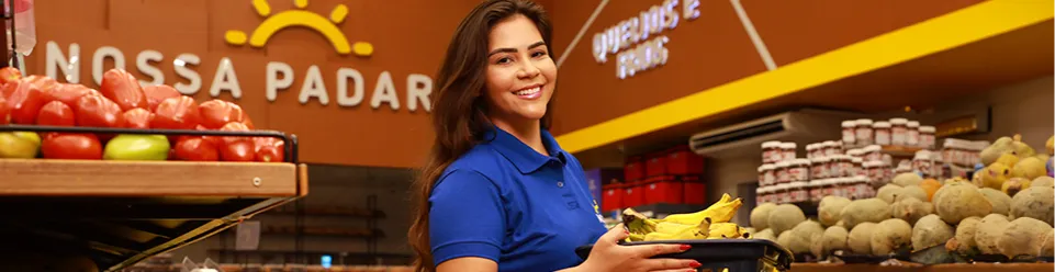 Funcionária sorridente, usando camisa azul, segurando uma cesta com bananas na seção de frutas e legumes, com fundo de padaria e queijos.