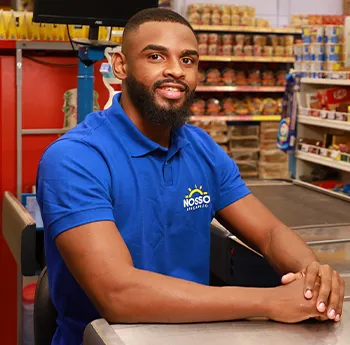 Um homem com barba, sorridente, veste camiseta polo azul com o logo do Nosso Atacarejo, sentado no caixa de supermercado, com os braços sobre o balcão.