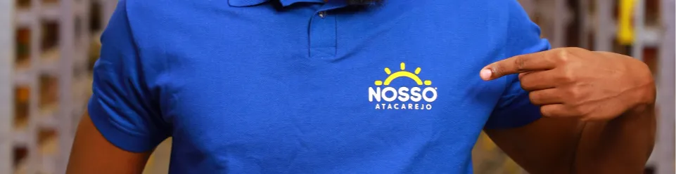 Colaborador usando camisa polo azul apontando para o logo “Nosso Atacarejo” pintada, com destaque para o logotipo (sol amarelo e texto branco).