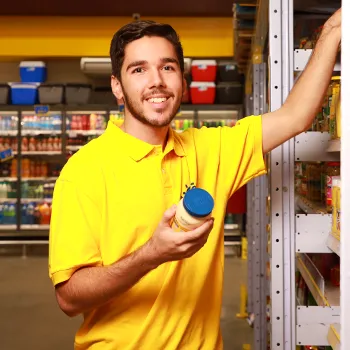 Um jovem sorridente, usando camiseta polo amarela, está organizando produtos em uma prateleira de supermercado e segurando um pote na mão.