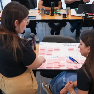 Duas colaboradoras sentadas em uma sala de reunião organizam ideias em uma folha grande sobre a mesa, utilizando vários post-its rosa. Ao redor, há garrafas, canecas e materiais de trabalho, indicando uma dinâmica colaborativa de planejamento.