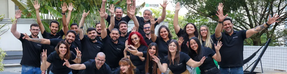 Grupo grande de colaboradores ao ar livre, sorrindo e com as mãos levantadas, transmitindo entusiasmo e união.