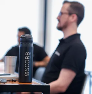 Garrafa térmica preta com o logotipo da SCORB em destaque sobre uma mesa. Ao fundo, desfocados, colaboradores participam de uma reunião ou treinamento, reforçando o ambiente corporativo.