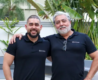 Dois colaboradores homens, usando camisetas pretas com o logotipo da SCORB, posam lado a lado em área externa com plantas ao fundo. Ambos estão sorrindo, com postura descontraída; um deles apoia o braço sobre o ombro do outro, demonstrando proximidade e parceria.