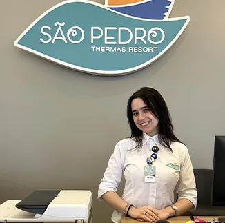 Recepcionista do Resort, com uma camisa branca como uniforme com o logo do Resort ao fundo e sorrindo.