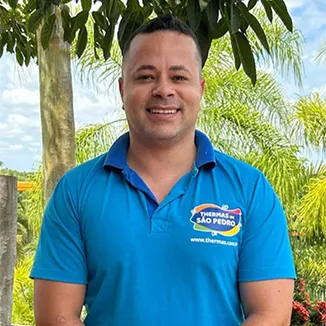 Colaborador do Thermas Parque, com uma camisa polo azul royal com o logo do Thermas ao lado direito. Ao fundo vegetação com coqueiros e árvores nativas.