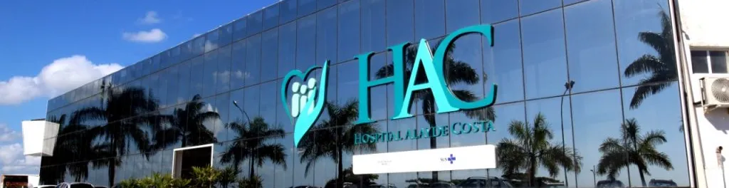 Fachada moderna do Hospital Alayde Costa (INAC), com prédio envidraçado, logotipo em destaque na cor azul e reflexo de palmeiras na estrutura, representando uma unidade hospitalar especializada em saúde.