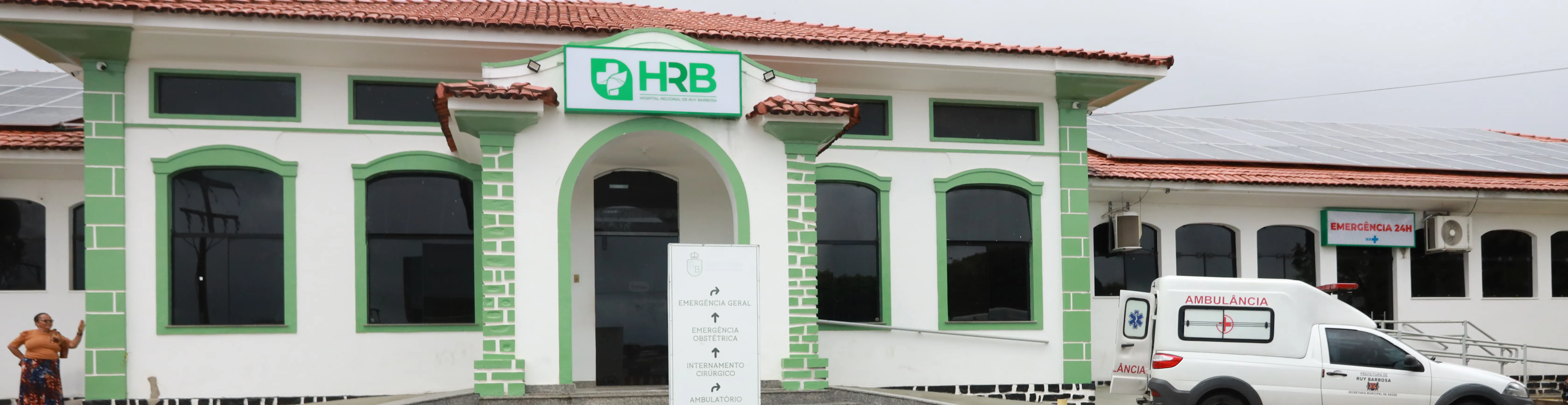 Fachada do Hospital Regional de Ruy Barbosa (HRB), com prédio histórico em tons de branco e verde, entrada central com arco, placa de identificação do hospital e ambulância estacionada ao lado, indicando atendimento de emergência.