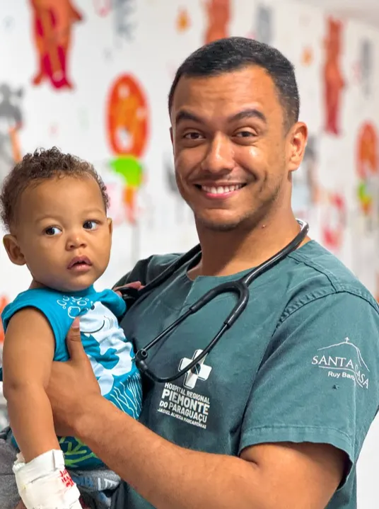 Médico do Hospital Regional Piemonte do Paraguaçu segurando um bebê em ambiente assistencial, com expressão de acolhimento e cuidado, reforçando o atendimento humanizado e a dedicação das equipes à saúde materno-infantil.