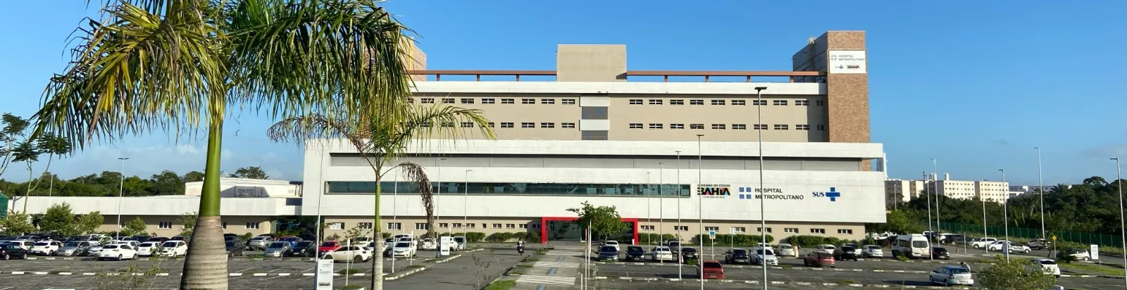 Fachada do Hospital Metropolitano, em Lauro de Freitas (BA), com prédio de grande porte, estrutura moderna, estacionamento em frente, palmeiras no entorno e identificação como unidade 100% SUS do Governo do Estado da Bahia.