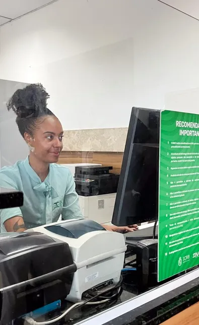 Recepcionista do INAC, sorridente e atenta, atrás de um balcão de atendimento, usando uniforme verde-claro, olhando para um monitor de computador; à frente há equipamentos de impressão e uma placa verde com recomendações.