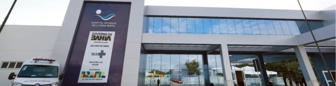 Fachada do Hospital Estadual do Litoral Norte, em Alagoinhas (BA), com estrutura ampla, fachada espelhada, ambulância posicionada na entrada e identificada como unidade SUS do Governo do Estado da Bahia.