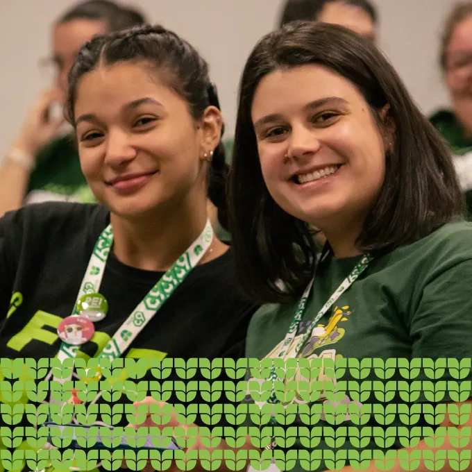 Uma fotografia de plano médio de duas mulheres jovens sorrindo para a câmera. Elas estão sentadas próximas uma da outra. A mulher à esquerda tem cabelos pretos e trançados e usa uma camiseta preta. A mulher à direita tem cabelos pretos e curtos e usa uma camiseta verde-escura. Ambas usam lanyards brancas com o logo da Univille. No fundo desfocado, há outras pessoas. Na parte inferior da imagem, há um padrão gráfico repetitivo de pequenas formas da logo da Univille em tons de verde claro.