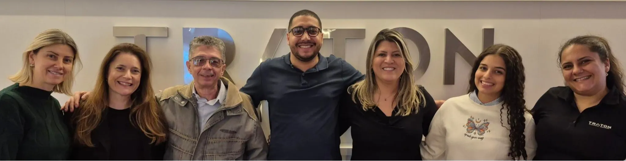 Sete pessoas posam sorrindo lado a lado em frente a uma parede com o logotipo da TRATON Financial Services. Elas demonstram proximidade e alegria, transmitindo o espírito de equipe e colaboração da empresa.