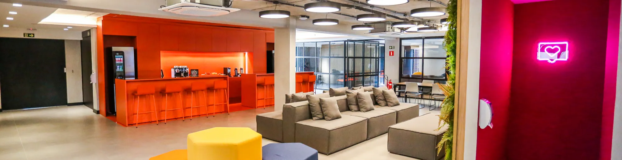 Ambiente interno do escritório da Watch Brasil de Curitiba. O espaço é amplo, com piso claro, teto aparente em concreto e luminárias circulares no teto. À esquerda, há uma área de copa na cor laranja, com balcão e banquetas altas. No centro, sofás cinza e pufes hexagonais nas cores laranja, amarelo, roxo e azul. Ao fundo, existem salas de reunião com paredes de vidro e mesas. À direita, há a entrada de um banheiro com parede rosa e um símbolo luminoso de coração.